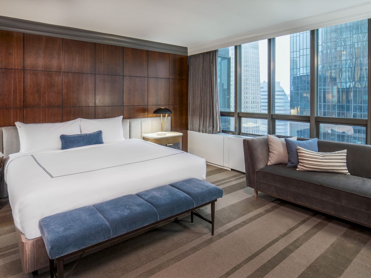 A modern hotel room with a large bed, blue bench, gray sofa, and city view through large windows. Decor includes wood paneling and neutral tones.