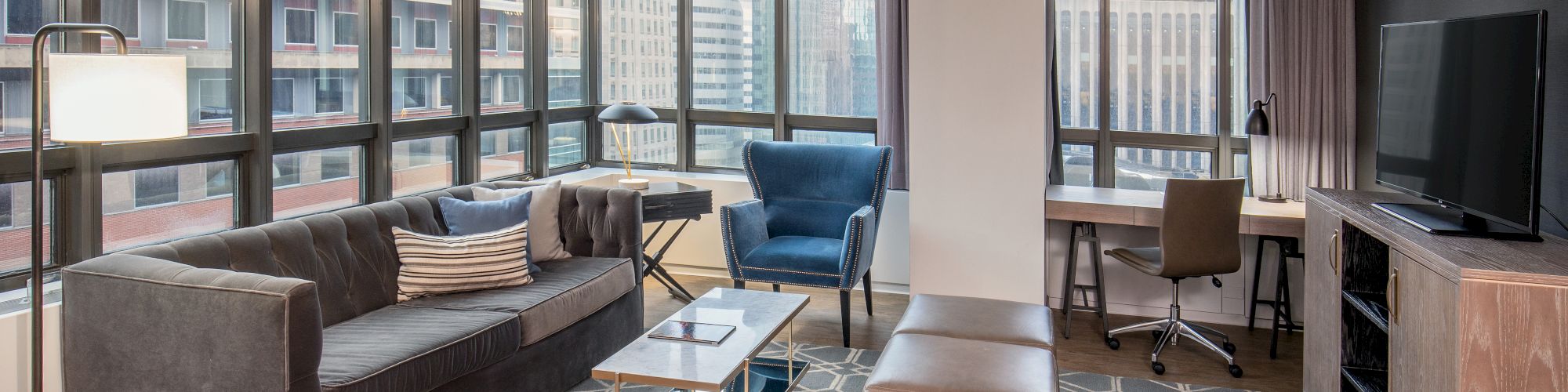 A modern living room with large windows, a sofa, chairs, TV, coffee table, desk, and urban view.