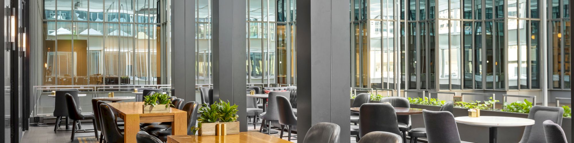 The image shows a modern indoor dining area with wooden tables, black chairs, and large windows offering a view of an office building.