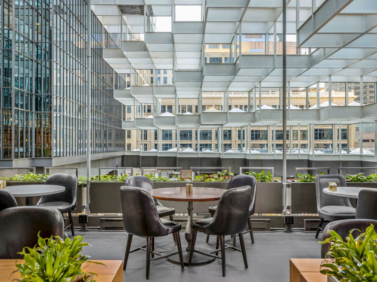 A modern outdoor dining area with round tables and black chairs, surrounded by plants, set within a high-rise building complex.