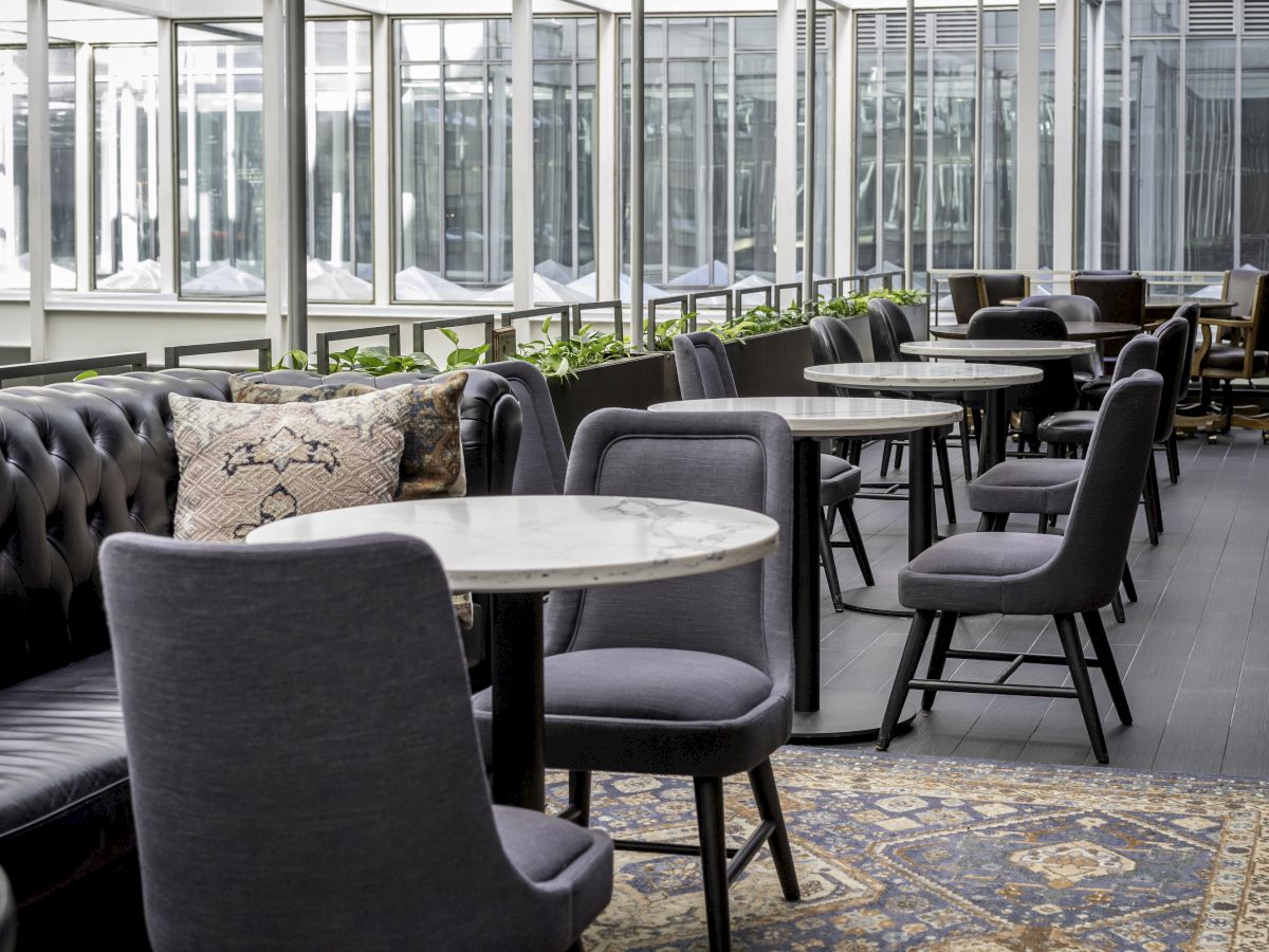 A modern indoor café with a mix of upholstered chairs and tufted bench seating, marble-topped tables, and large windows.