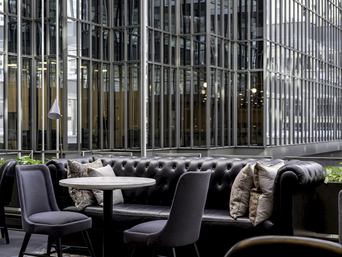 The image shows an outdoor seating area with a black tufted couch, gray chairs, a round table, and a modern glass building in the background.