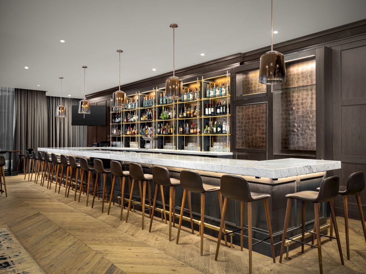 A modern bar with a marble countertop, lined with wooden stools, shelves stocked with bottles, and pendant lighting fixtures hanging from the ceiling.