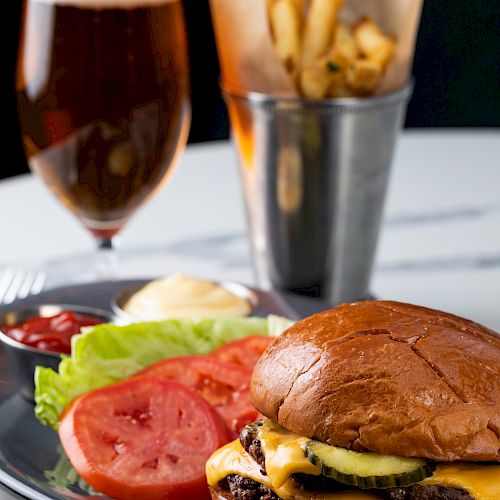 A cheeseburger with lettuce and tomato on a plate, accompanied by fries in a metal container and a glass of beer in the background.