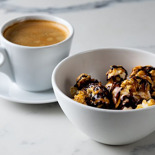 A cup of coffee sits next to a white bowl of caramel and chocolate drizzled popcorn on a marble surface.