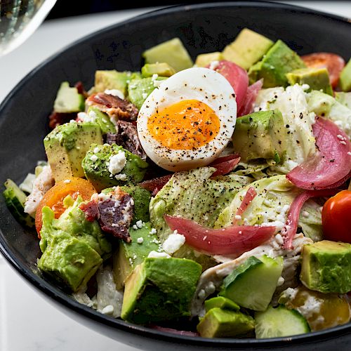 A fresh salad with avocado, cherry tomatoes, red onions, hard-boiled egg, and various greens, served in a black bowl with a glass of white wine.