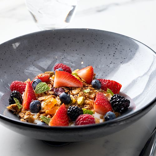 A grey bowl filled with fresh berries, granola, and yogurt on a marble surface, accompanied by a knife and fork, with a glass in the background.