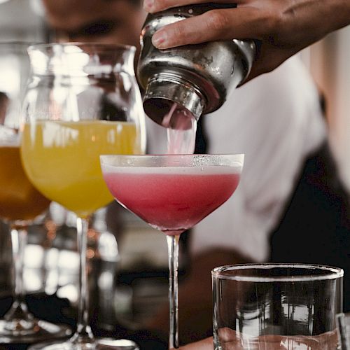 A bartender is pouring a pink cocktail from a shaker into a glass, with two other colorful drinks in the background.