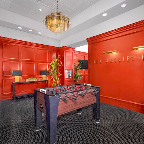 A room with red walls features a foosball table, plants, a chandelier, and a sign that reads 