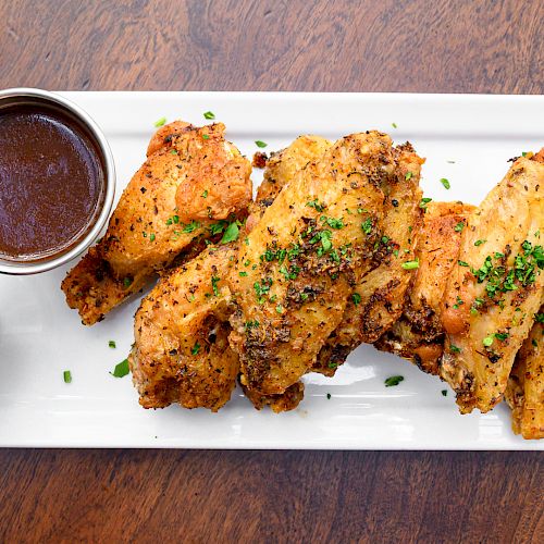 The image shows a rectangular white plate with seasoned chicken wings, garnished with green herbs, accompanied by two dipping sauces.