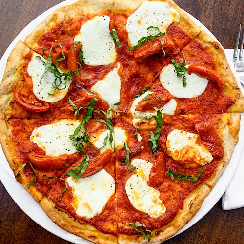 The image shows a margherita pizza topped with fresh mozzarella, tomato slices, and basil, served on a wooden table with a fork and knife on a napkin.