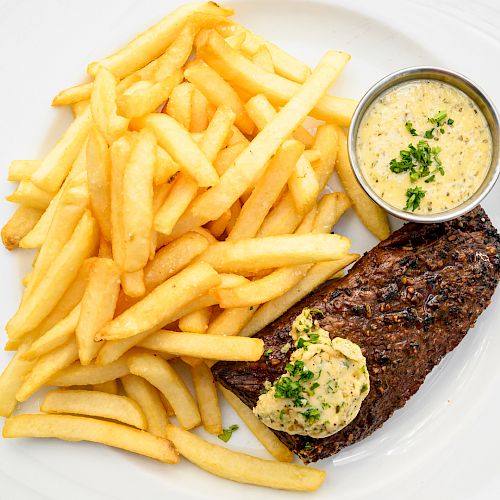 A plate of French fries and a grilled steak topped with herb butter, accompanied by a cup of sauce, utensils placed on the table.