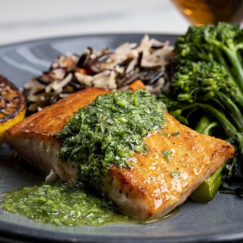 A plate of grilled salmon topped with green sauce, served with wild rice, broccolini, and a grilled lemon slice on the side, is presented.