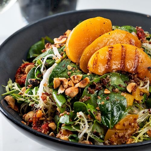 A fresh salad with grilled peach slices, leafy greens, quinoa, and chopped nuts is in a black bowl on a table with a glass of white wine.