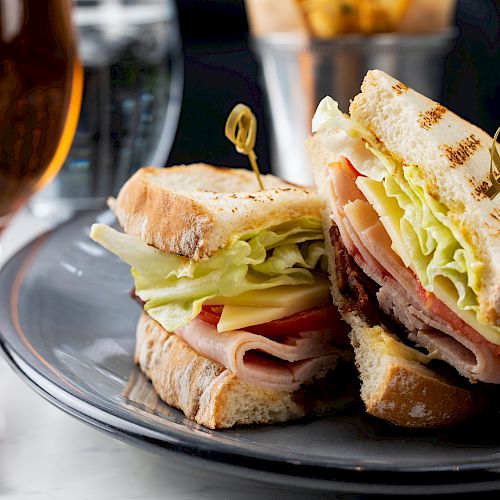 A sandwich with lettuce, cheese, tomato, and meat on a plate, served with fries, a glass of beer, and a fork on the side of the table.