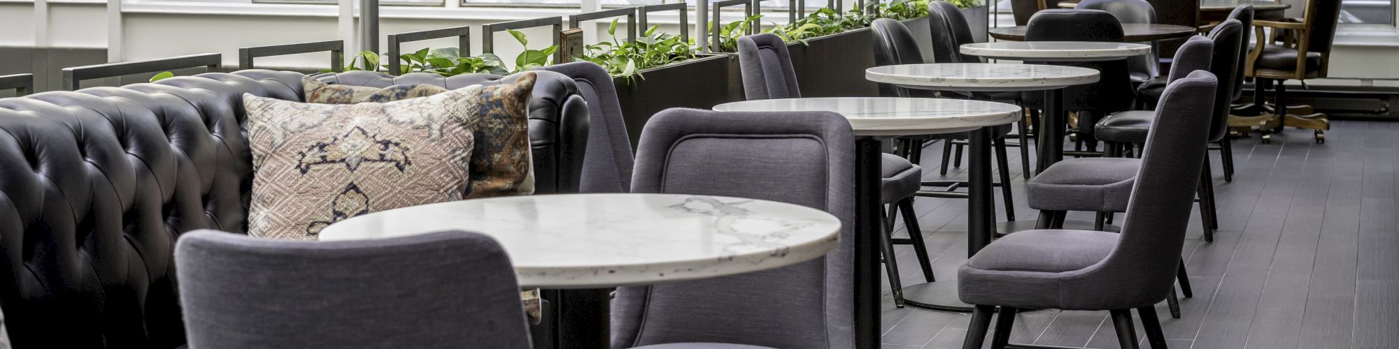 A modern indoor café with round marble tables, cushioned chairs, and a tufted bench alongside a wall of windows, well-lit and stylishly decorated.