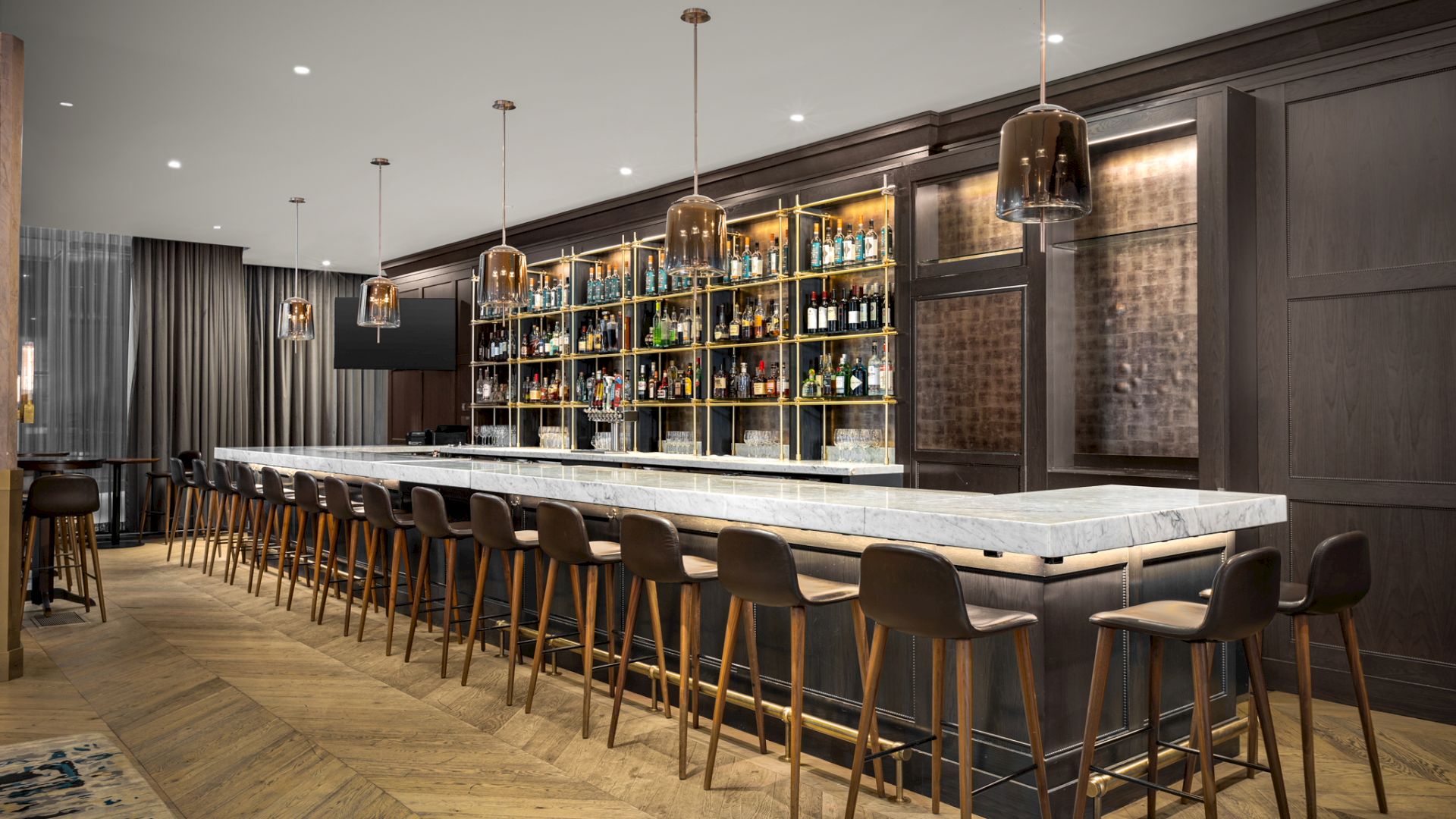 This image shows a modern, stylish bar with a long marble counter, wooden bar stools, pendant lights, and shelves stocked with various bottles.