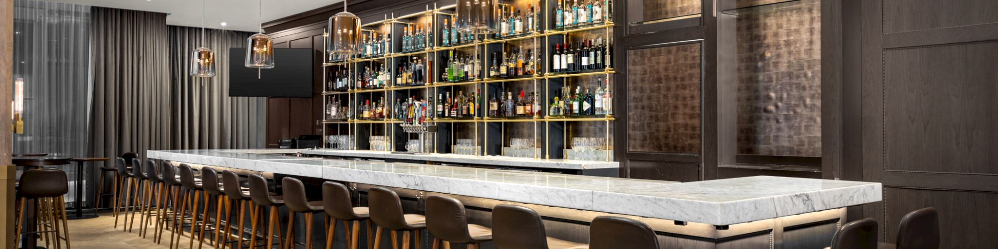 This image shows a modern, stylish bar with a long marble counter, wooden bar stools, pendant lights, and shelves stocked with various bottles.