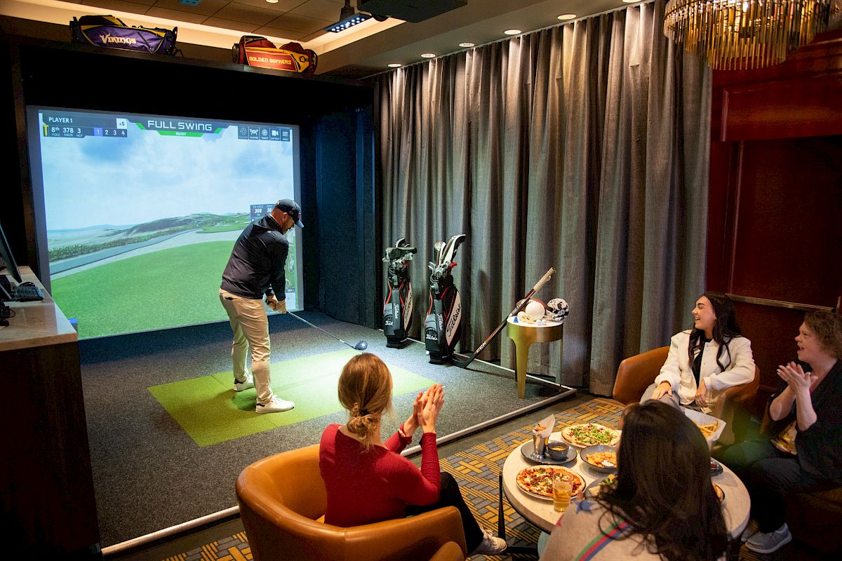 People enjoying an indoor golf simulator; one is playing while others watch and clap, surrounded by golf accessories and food.
