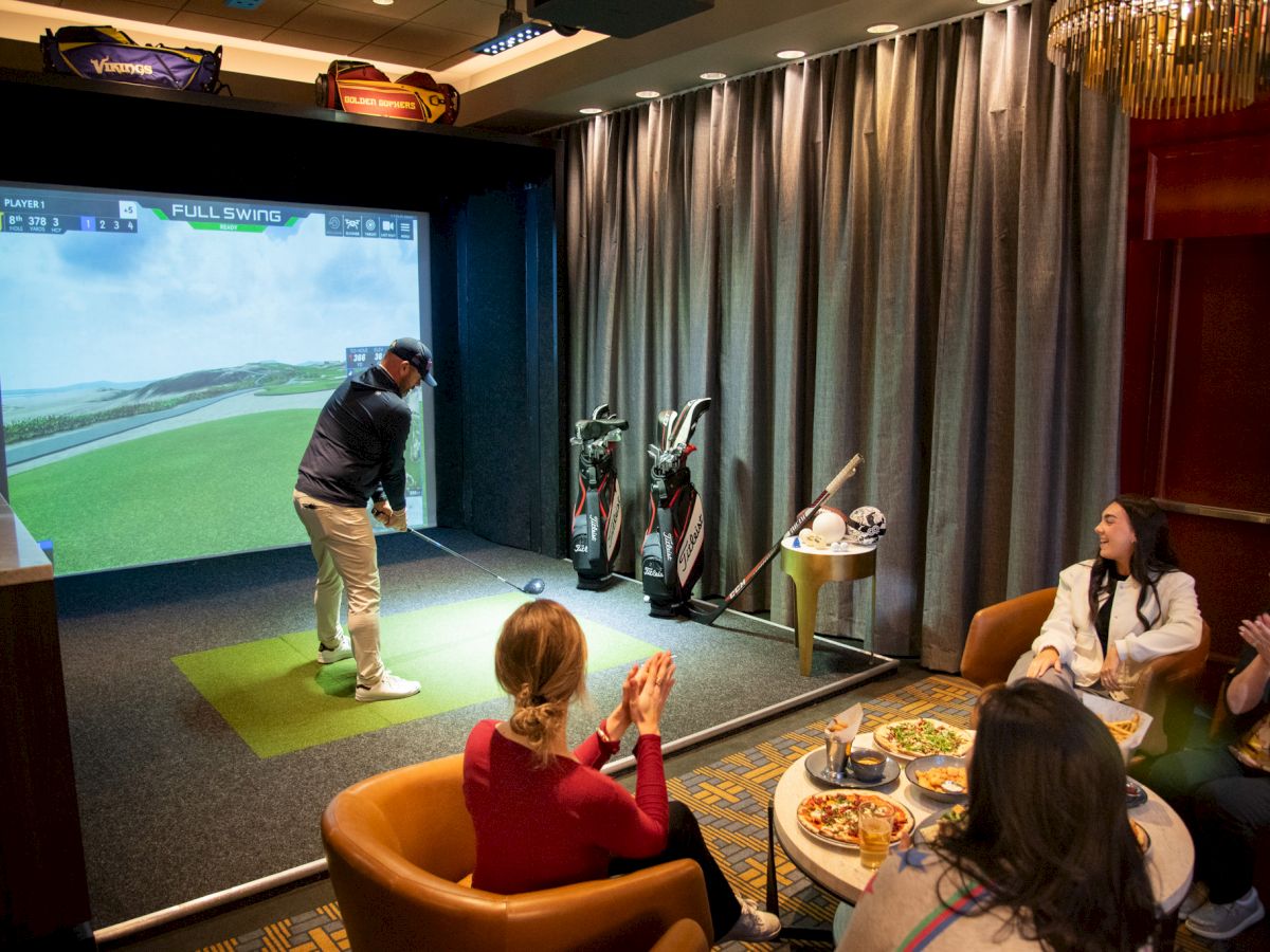 People enjoying an indoor golf simulator; one is playing while others watch and clap, surrounded by golf accessories and food.