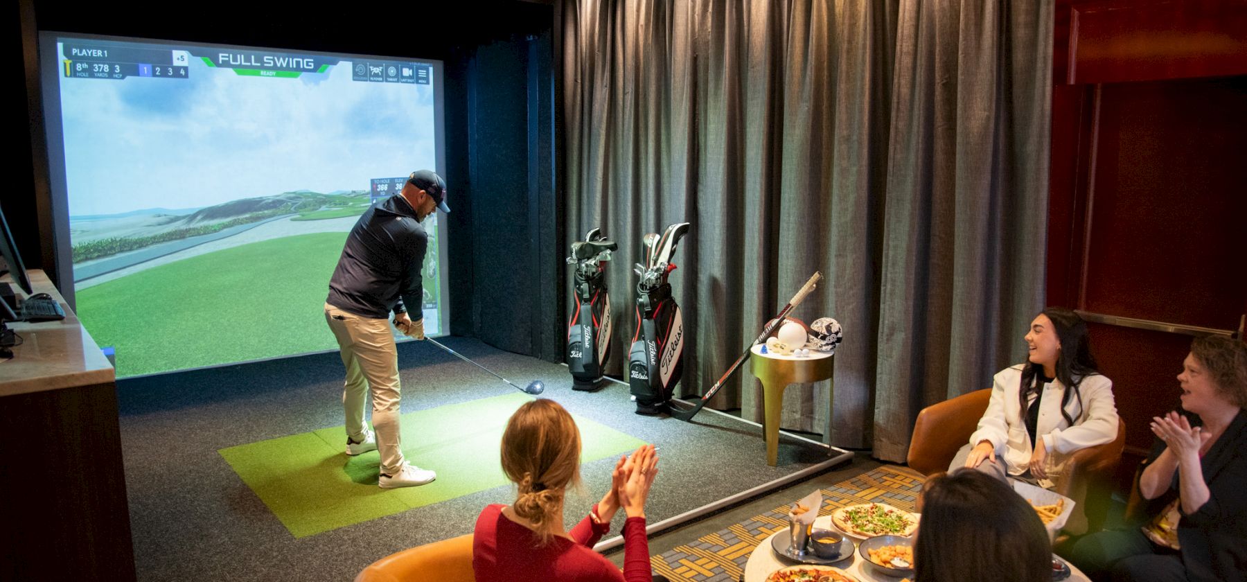 People enjoying an indoor golf simulator; one is playing while others watch and clap, surrounded by golf accessories and food.