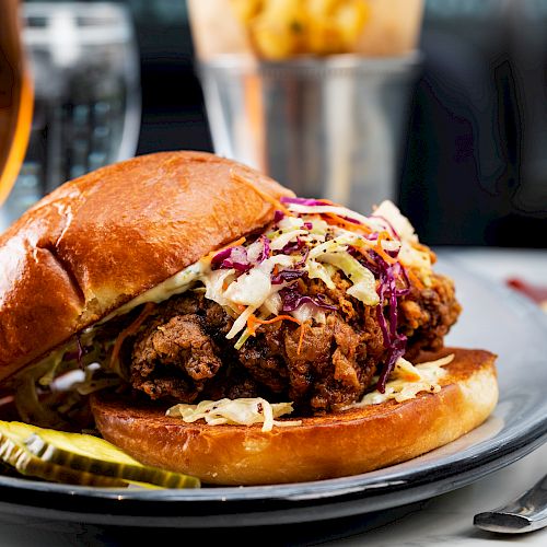 A fried chicken sandwich with coleslaw and pickles on a bun, served on a plate with a side of sauce, and a drink in the background.