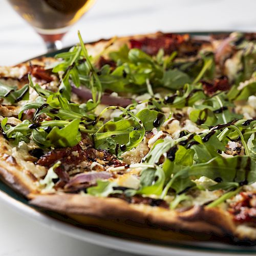 The image shows a delicious pizza topped with arugula, red onions, sun-dried tomatoes, and a drizzle of balsamic glaze on a thin crust.