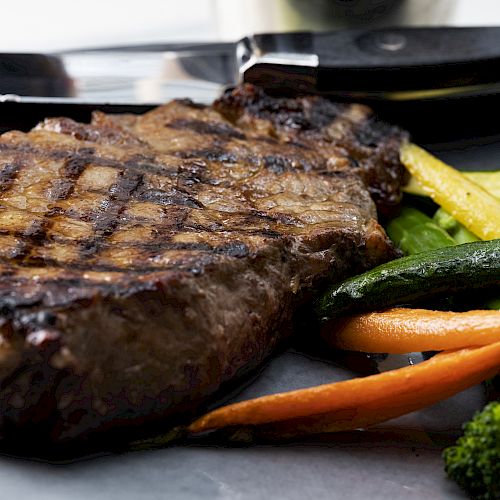 A grilled steak is served with mixed vegetables, including broccoli, carrots, and zucchini, and a knife is placed on the side of the plate.