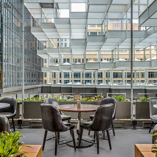 A modern outdoor seating area with round tables, black chairs, green plants, and a grid-like ceiling, surrounded by tall glass buildings and city views.