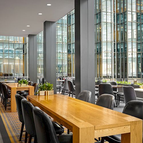 A modern indoor seating area with wooden tables, black chairs, and potted plants beside large glass windows in a high-rise building.