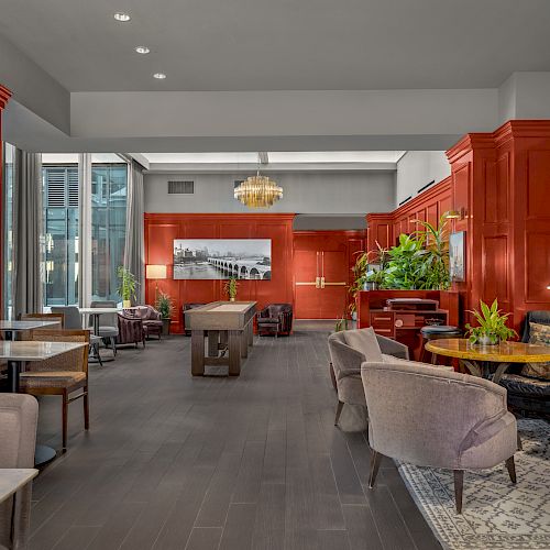 The image shows a stylish lounge area with red paneled walls, various seating, tables, and a pool table in the center, with plants and decor.