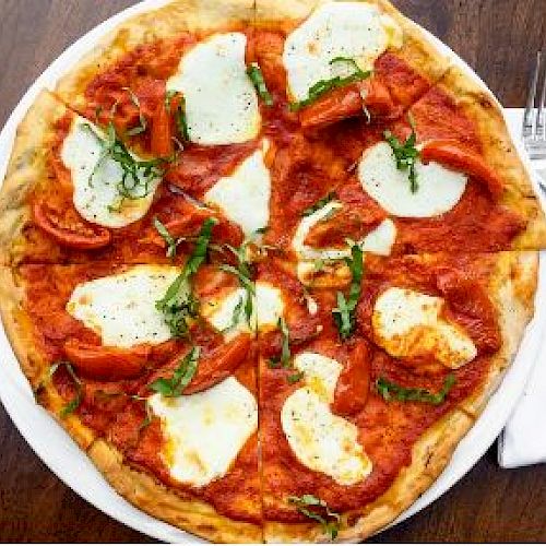 A Margherita pizza topped with mozzarella cheese, tomato sauce, and basil leaves, served on a white plate next to a napkin with a fork and knife.