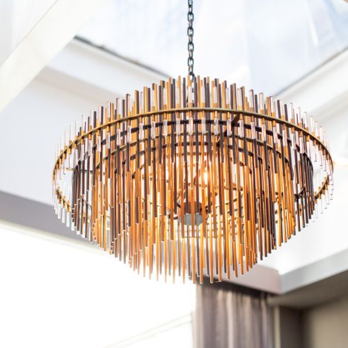 A modern chandelier with multiple vertical rods hanging from a ceiling, illuminated warmly in a bright room with large windows.