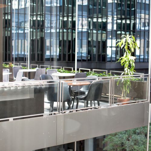 The image shows an outdoor terrace with tables and chairs, set against a backdrop of modern glass buildings and some plants for decoration.