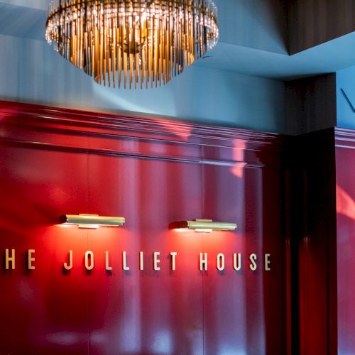 The image shows a red wall with the text "THE JOLLIET HOUSE" and two lights beneath a decorative chandelier.