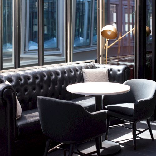 A stylish seating area with a black leather sofa, two chairs, a round table, and a modern lamp by large windows.