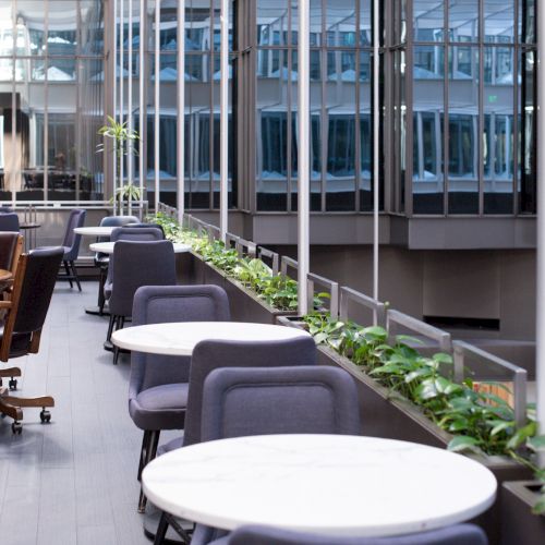 The image shows a modern indoor workspace with round tables, office chairs, and greenery along a glass wall, creating a bright atmosphere.