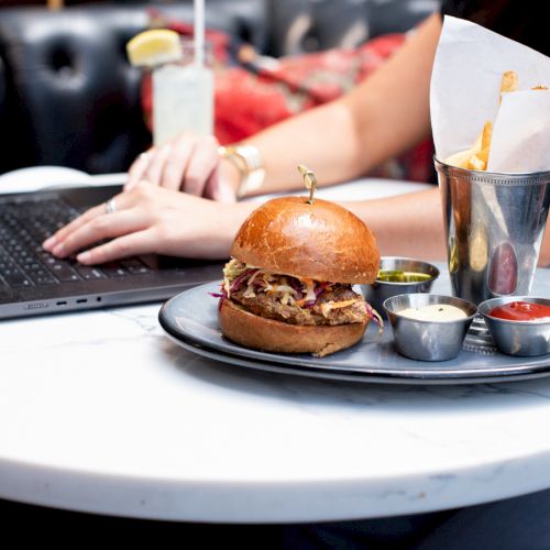 A person using a laptop at a table with a burger, fries, and dipping sauces on a plate.