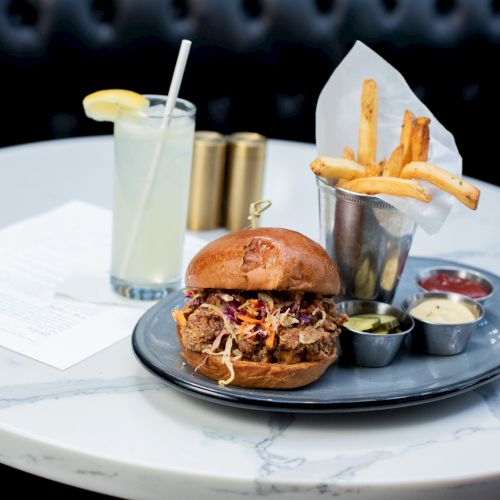 A burger with coleslaw, fries in a cup, and dipping sauces on a plate next to a lemonade on a marble table.