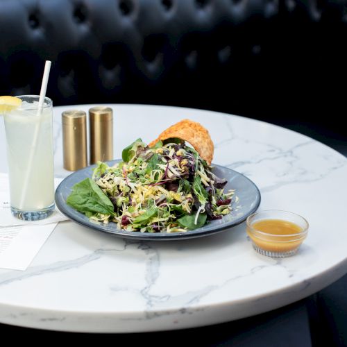 A fresh salad with greens, a drink with a lemon wedge, a dressing, and a menu sit on a marble table.