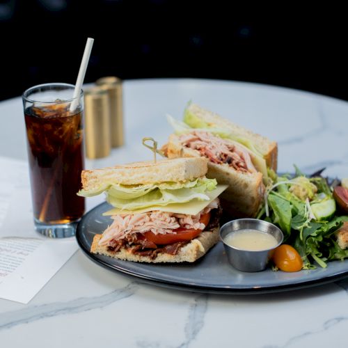 A sandwich with turkey, lettuce, and tomato, a side salad, a small sauce dish, and a glass of soda with a straw on a table.