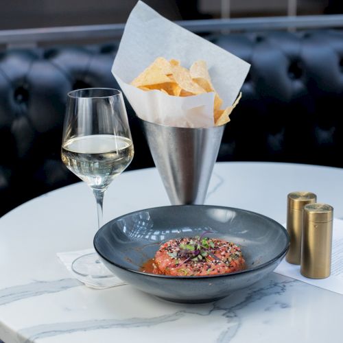 A white wine glass, a bowl with a finely prepared dish, and a cone of chips are on a marble table with a dark cushioned background.