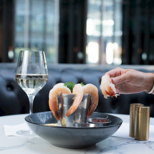 A person is taking a shrimp from a dish, accompanied by a glass of white wine, on a marble table in a modern dining setting.