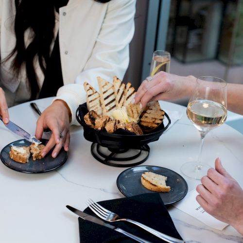 Two people enjoy bread with butter and wine at a table. The setting includes a basket of grilled bread and glasses of white wine.