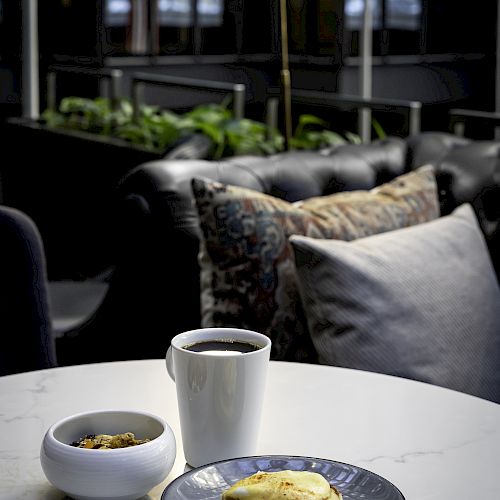 A cozy table setup with a cup of coffee, a plate of pancakes, a bowl of granola, and a lamp illuminating the scene against a stylish couch.