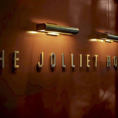 The image shows a wall with the text "THE JOLLIET HOUSE" illuminated by two wall-mounted lights above the letters, creating a warm ambiance.