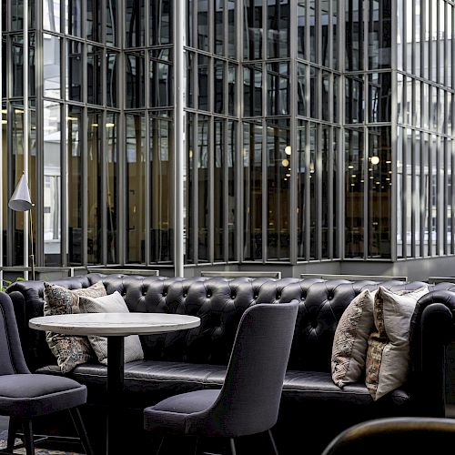 A modern, outdoor seating area with a black leather sofa, gray chairs, and a round table, set against a backdrop of reflective glass buildings.