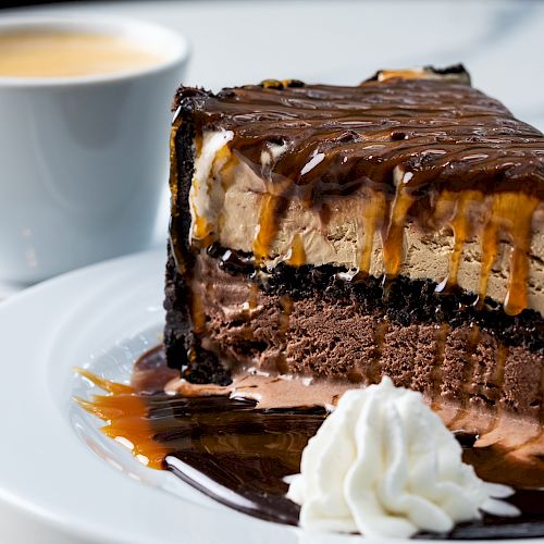 The image shows a slice of layered chocolate cake with whipped cream on a white plate, and a cup of coffee in the background, ending the sentence.