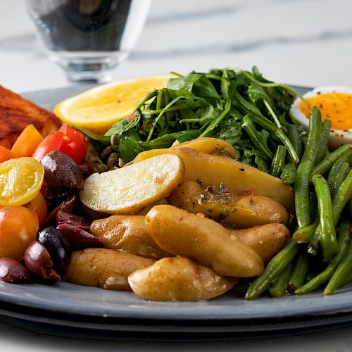 A plate with baked salmon, potatoes, green beans, cherry tomatoes, baby arugula, olives, a boiled egg, and pickled vegetables.