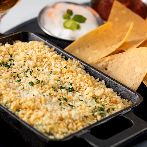 The image shows a dish, possibly a casserole or dip, topped with breadcrumbs and herbs, accompanied by tortilla chips and a side of dip or sour cream.